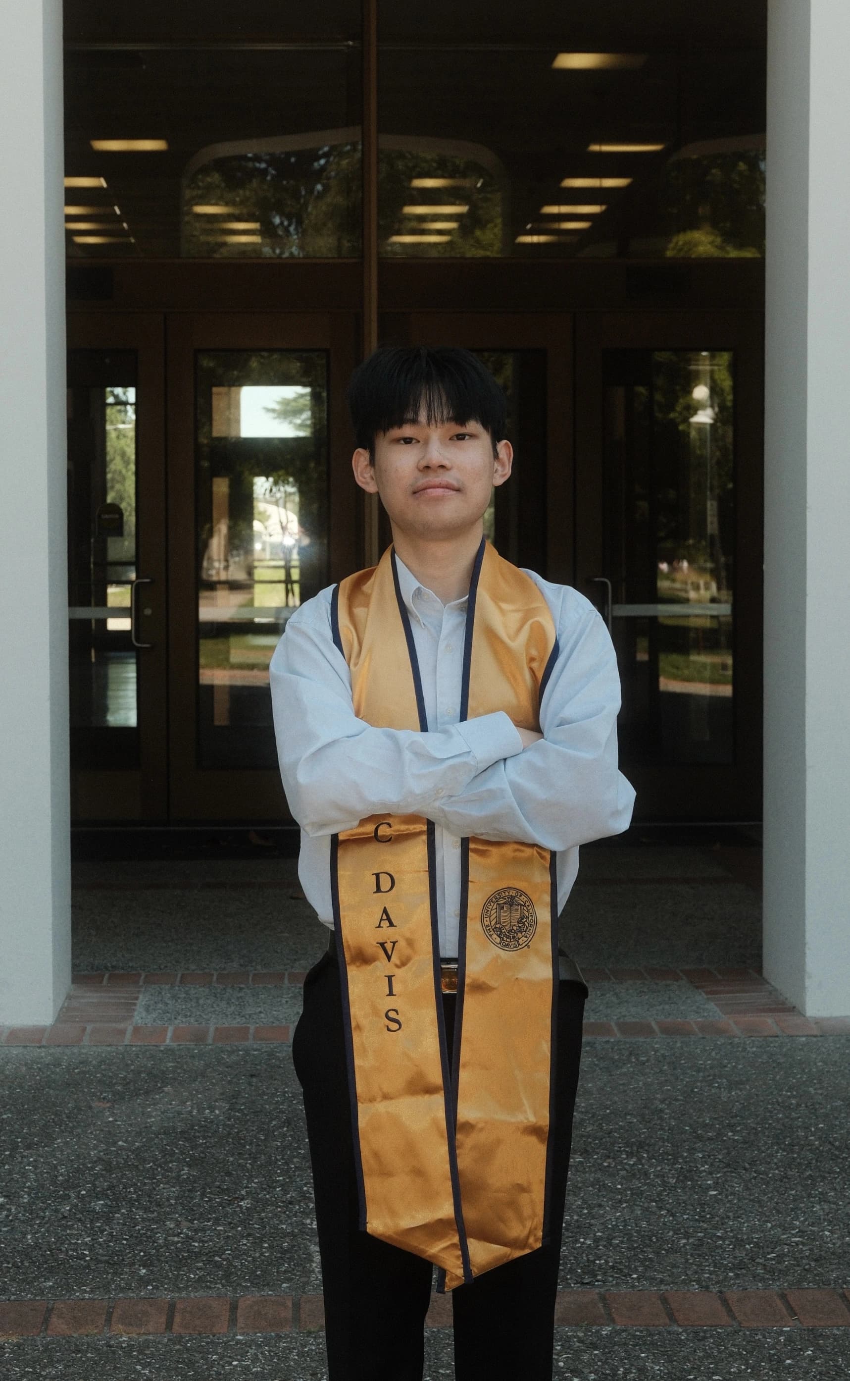 Tony Lin standing with arms crossed in a light blue shirt and a gold graduation stole